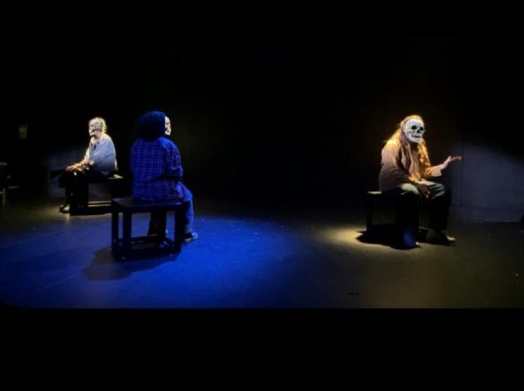 Three actors sit on blocks in their own pool of a colored spotlight. One is gesturing. They all have skull emojis covering their faces.