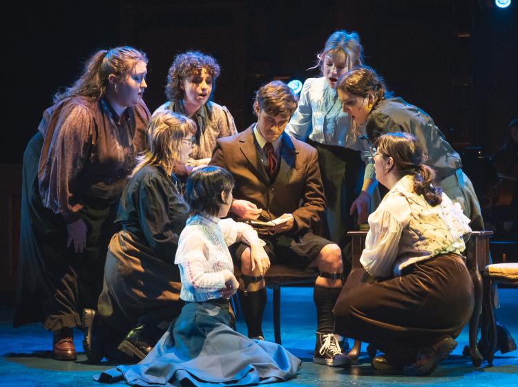 A young man sitting and writing in a journal, with young women gathered around him reacting positively to what they see him writing.