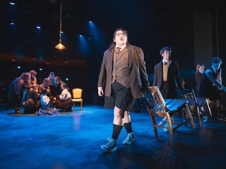 Young men singing and pulling chairs behind them in single file, and a group of young women gathered around a single young man who is sitting in a chair and writing in a journal.