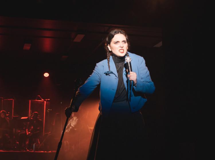 A young woman holding a mic and mic stand with an anguished expression as she sings. The onstage band can be seen behind her with dramatic red lighting.