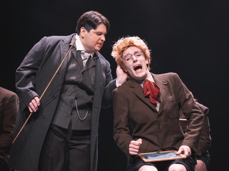 A teacher holding a stick and twisting the ear of a young man sitting in a chair whose face is in anguish. Both are wearing formal, boarding school clothing.