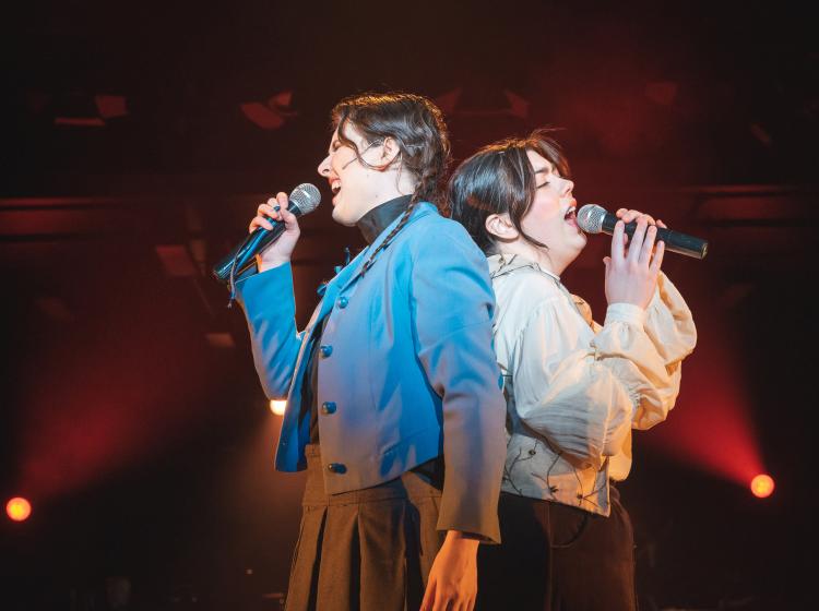 Two young women stand back to back singing passionately into microphones with dramatic lighting.