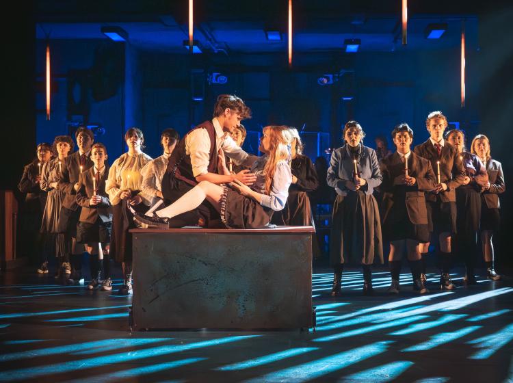 A young man and young woman on a stand in the act of sexual foreplay with a group of young people lined up behind them with candles singing.