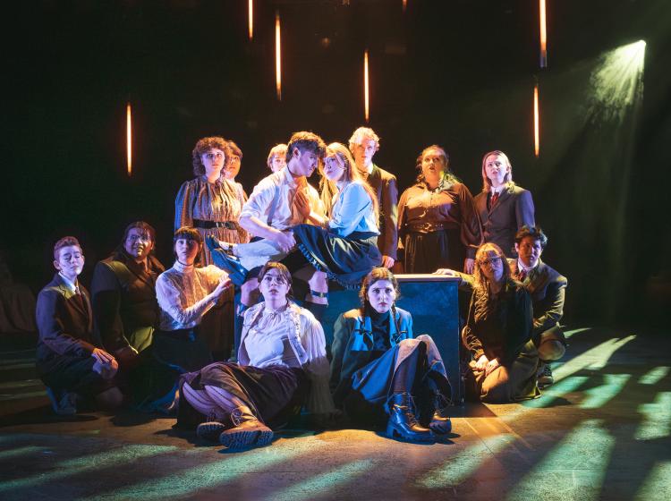 A bunch of young men and young women posed on a stage with a couple in the middle embracing. The lighting gives the sense of a hayloft. 