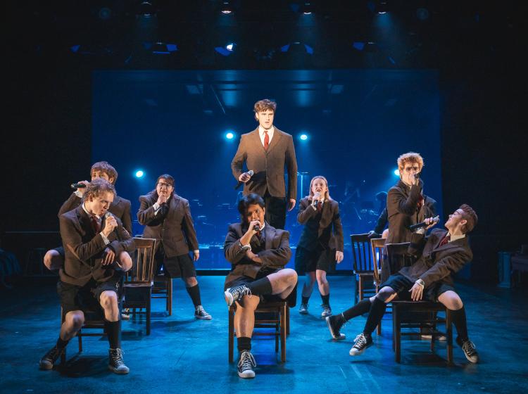Young men in boarding school uniforms rocking out on classroom chairs with dramatic lighting.