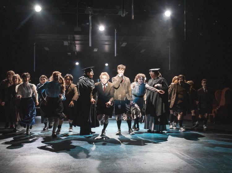 One young man singing in the center in a spotlight, while a line of young people are running intensely past the singer toward the audience. 