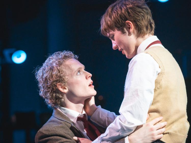 One young man straddles another young man and they embrace looking into each other's eyes.