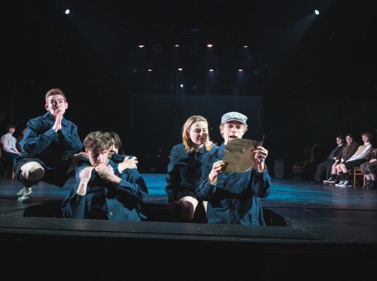 A bunch of young men are standing in a bit. One is reading a letter and the other boys have a second boy trapped who is struggling to escape.