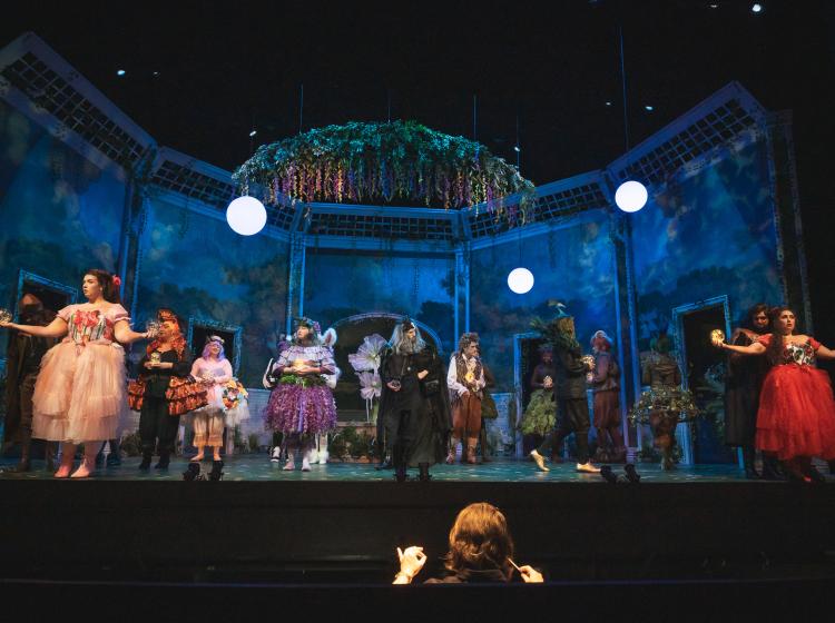 A wide angle photo of the Lincoln Performance Hall stage with the set of Cosi fan tutte and many actors in costume. The orchestra conductor can be seen in the pit.