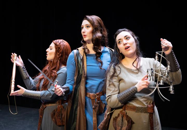 Three young women in rugged medieval costumes, carrying props including a sword, bells and mock flute