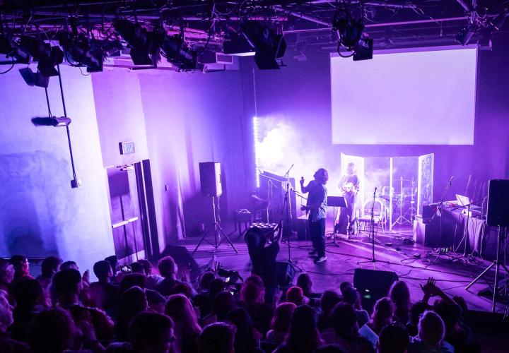 Audience members looking at a stage with purple lighting and musicians, and lighting hung in rafters overhead.