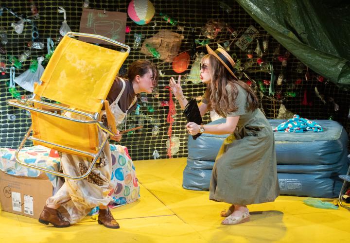 Actress one holding a yellow lawn chair. Actress two with her hand up like she is trying to hush actress one. From PSU Theater's "Frontieres sans Frontieres"