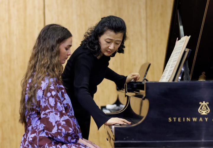 A teacher leaning over a piano demonstrating something while the student sits observing.