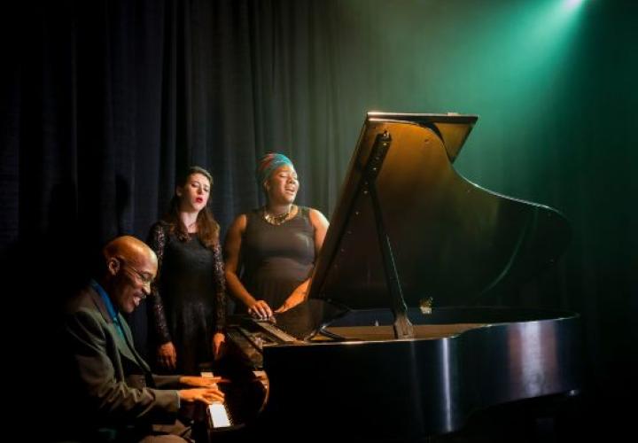 A man at the piano with two women singing beside him.