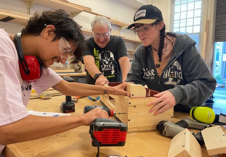 Two students and an adult mentor in PSU's Theater Arts set build shop using an electric screw driver to  hold pieces of wood together.