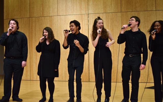 A group of singers in all black perform on a bright wooden stage