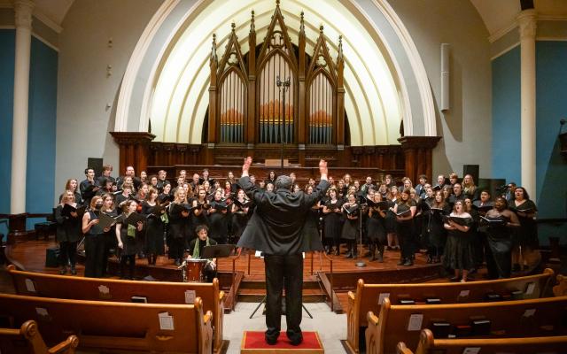 Conductor dramatically gesturing to a large choir in a church with an impressive pipe organ.