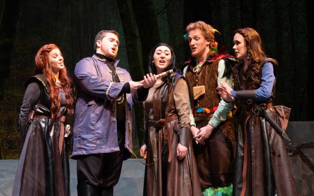Actors on stage in elaborate fantasy costumes sing while one holds a flute.