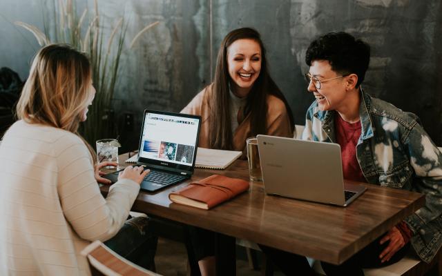 three people sitting at a table with their laptops working and laughing