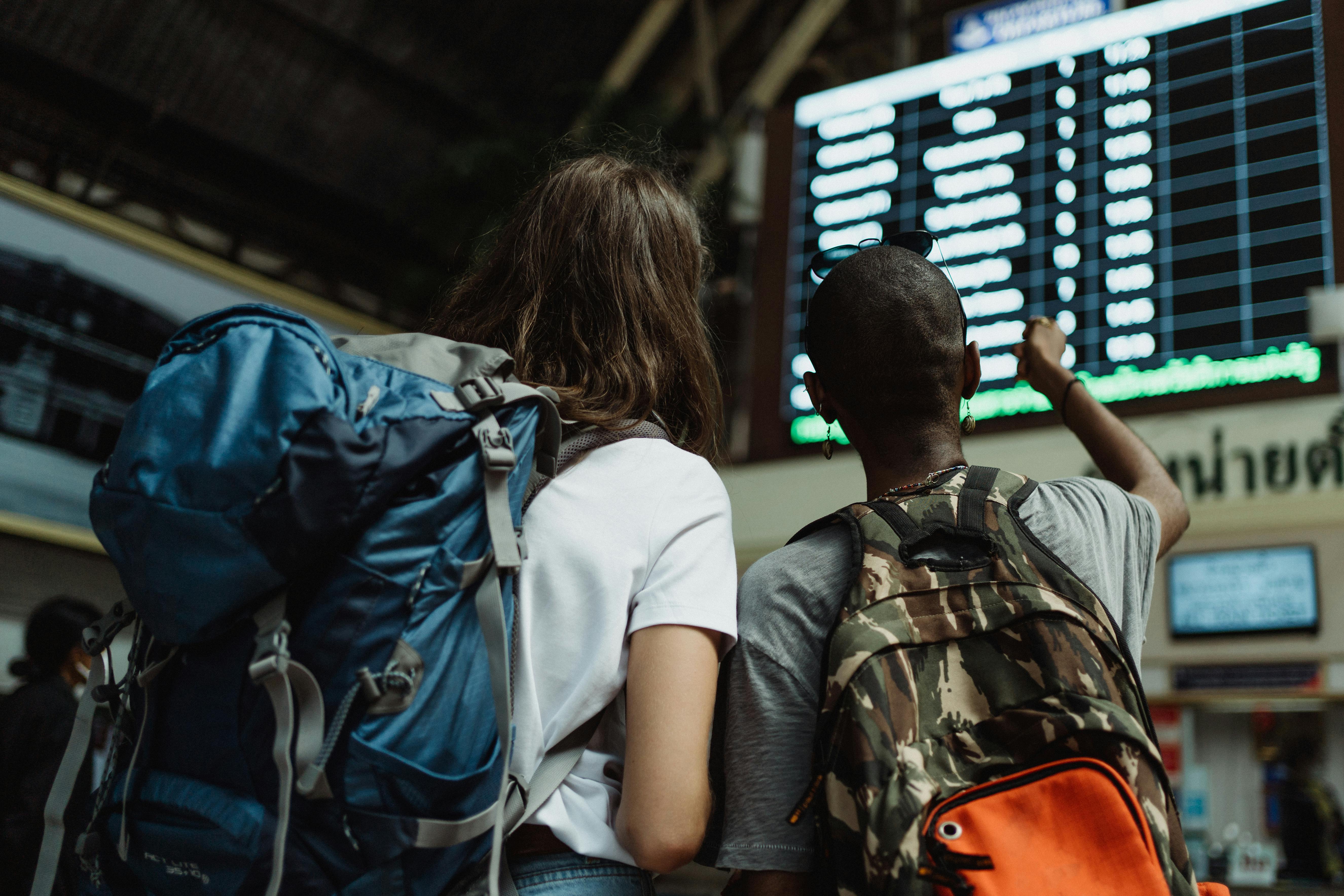 Two women wearing backpacks in a train station looking at departure times