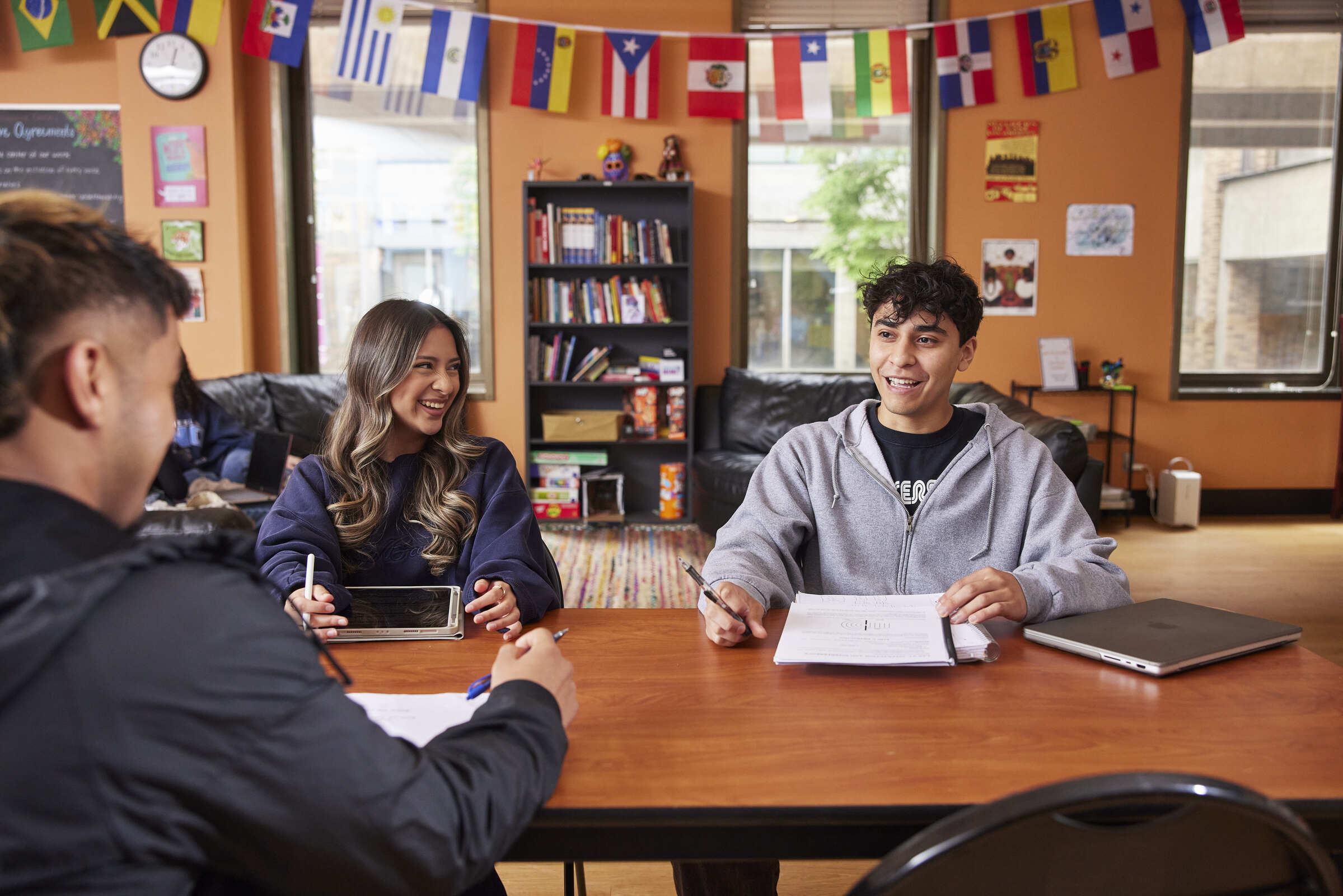 Three students laughing as they study together in a room adorned with international flags