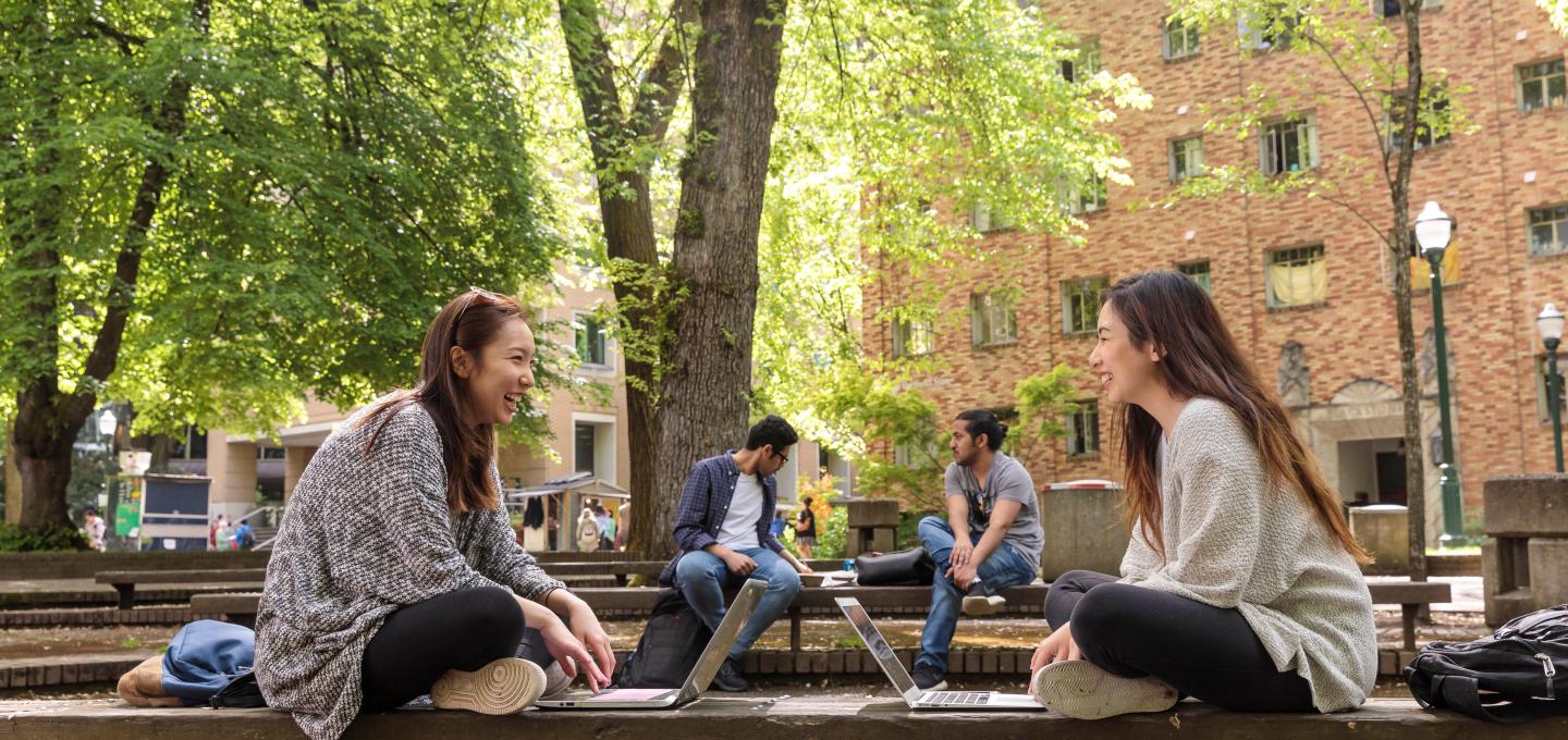 Two students sit facing each other while working on their laptops in the Park Blocks.