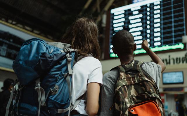 Two women wearing backpacks in a train station looking at departure times