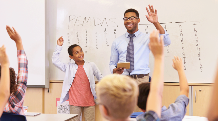 A Black male teacher in a light blue button-down and glasses stands in front of a whiteboard with a young student, both smiling with their hands raised.