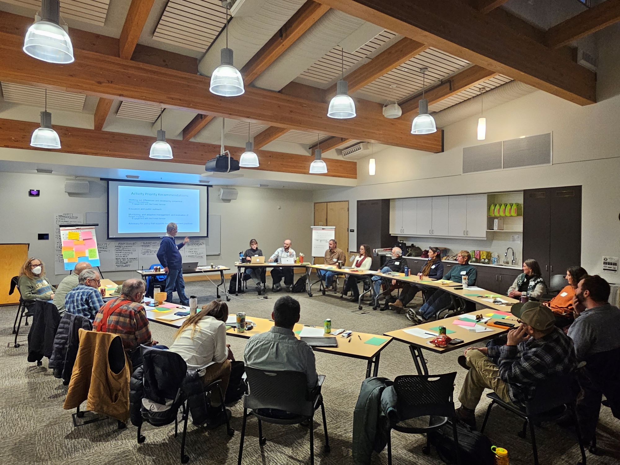 Group of people sitting in a classroom behind desks in a large circle, looking toward the front of the room where a person is presenting