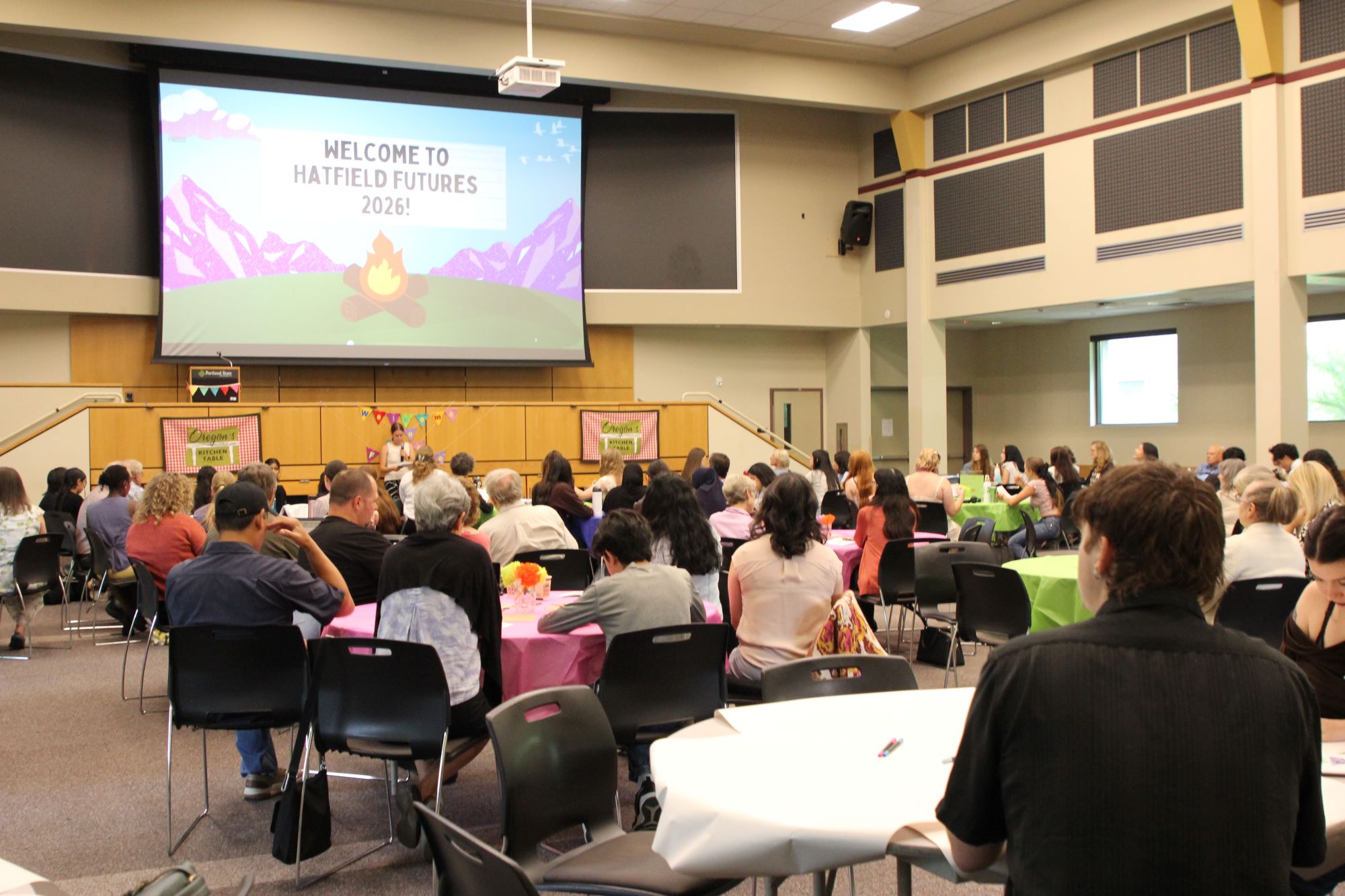 An audience of people sitting around round tables, looking forward toward the front of the room where a student is speaking. Behind the student is a presentation slide that reads, "Welcome to Hatfield Futures 2026!"