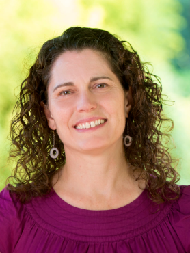 Tia is a white woman with brown curly hair. She is wearing a deep pink top and is looking at the camera, smiling.
