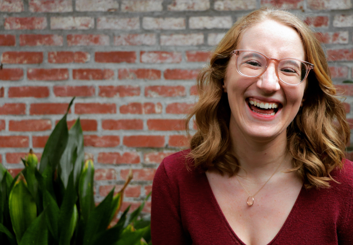 Caroline is a white woman with pink glasses and she is grinning at the camera. She is wearing a red sweater and is standing in front of a faded red brick wall