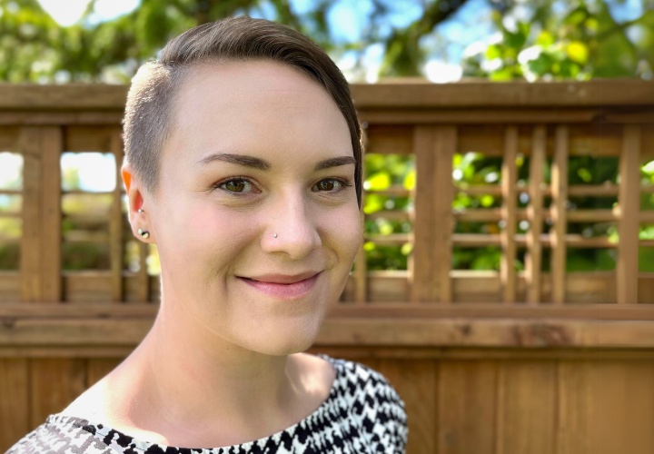 Erin is a white woman with brunette hair, which is partially shaved on one side and longer on the other. She is smiling at the camera, standing outside in front of a brown fence and foliage.