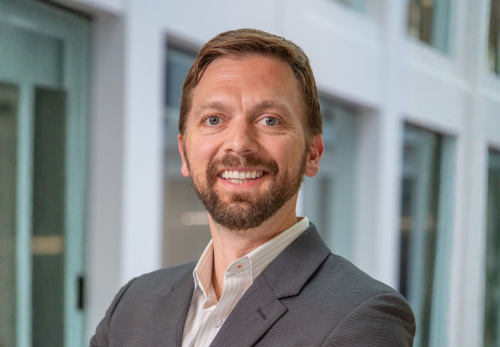 Jacen is a white man with brown hair and a beard. He is smiling at the camera in a gray suit.