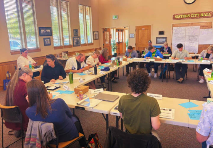City of Sisters Civic Leadership Academy participants sitting at tables in a square together. Some folks are seen talking to each other, others are looking down at the table in front of them.