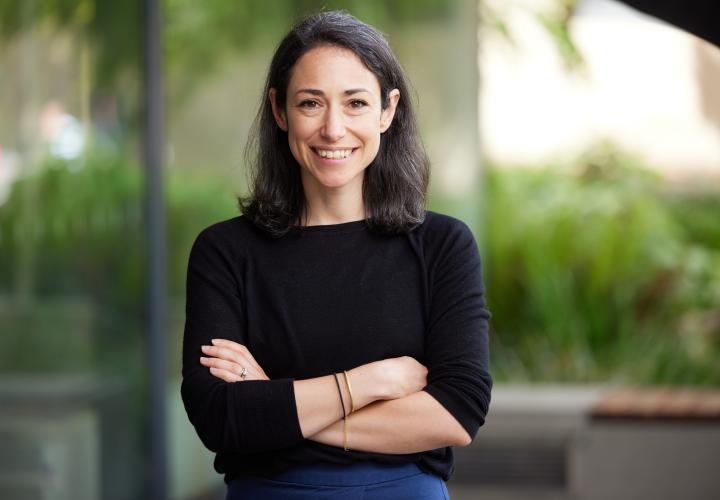A headshot of Abby Chroman. Abby has mid-length dark dark, and is wearing a black shirt. She is standing with her arms crossed, smiling at the camera.