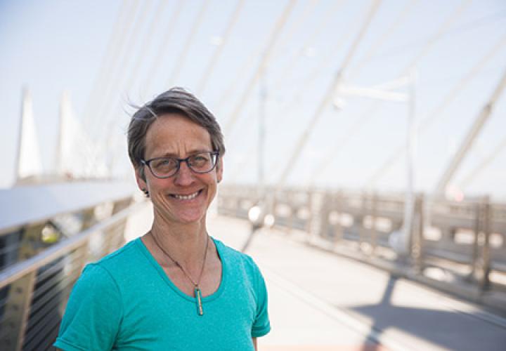 Jennifer is a white woman with short brown hair and glasses. She is standing on the Tilikum Bridge, smiling at the camera