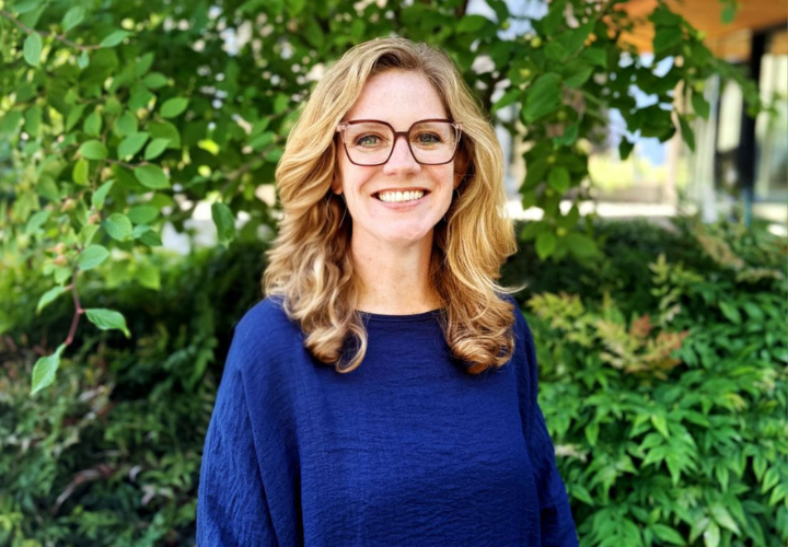 A headshot of Katie Pearmine, a woman with medium curly blonde hair. She is wearing a blue shirt and glasses and she is grinning at the camera