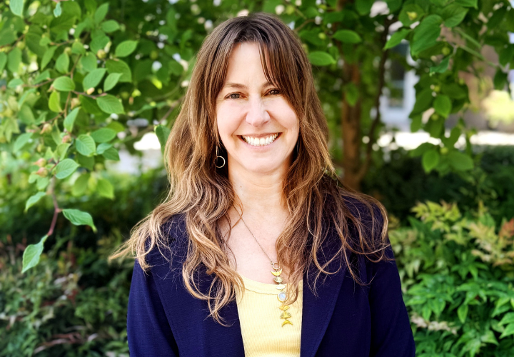 A headshot of Robin Harkless, a woman with long brown-copper hair. She is wearing a blue suit jacket and yellow shirt, with a long necklace. She is grinning at the camera.