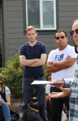 A group of people watching an older gentleman address them. The man is wearing a green and white plaid shirt and sunglasses, with his arms outstretched. Several others watch him in the background of the photo.