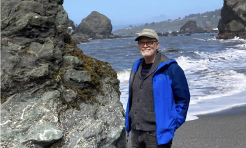 Steve Greenwood is standing on the beach, with waves crashing up behind him. He is wearing a blue jacket, tan baseball hat, and glasses. He is looking at the camera, smiling.