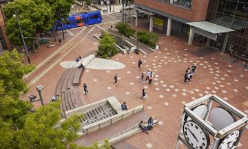 A dozen or so people milling about on the red brick Urban Plaza, photographed from above. 