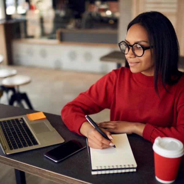 Woman writing on notebook with a laptop in from of them.