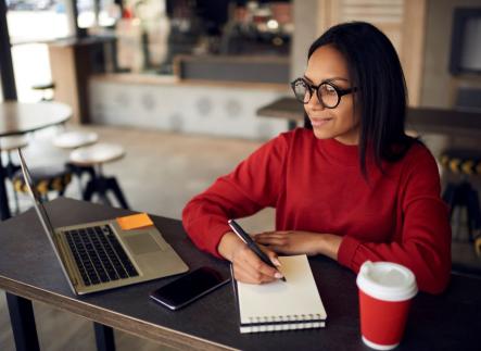 Woman writing on notebook with a laptop in from of them.