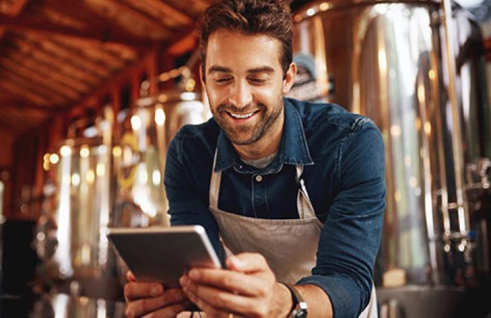 Man in apron smiling at tablet with brewery equipment behind him