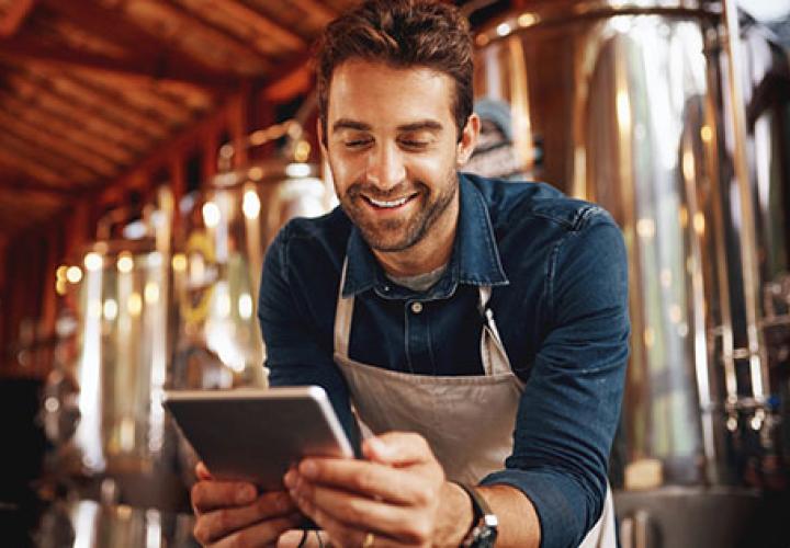 Man in apron smiling at tablet with brewery equipment behind him