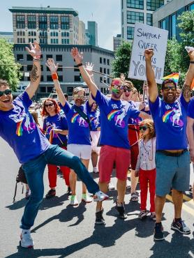 a group of people all wearing purple QRC shirts with a unicorn puking a rainbow stand outside happily with their arms raised in celebration at pride