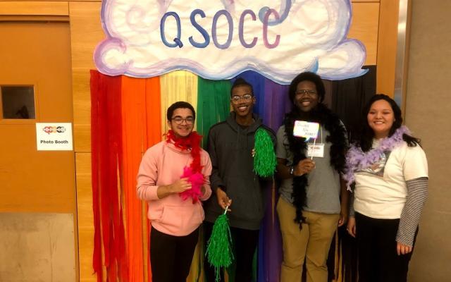 4 QTBIPOC stand in front of a cloudy rainbow QSOCC sign