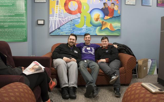 3 white queers sit on a couch in the qrc with their arms around each other, all smiling, udnerneath a QRC mural