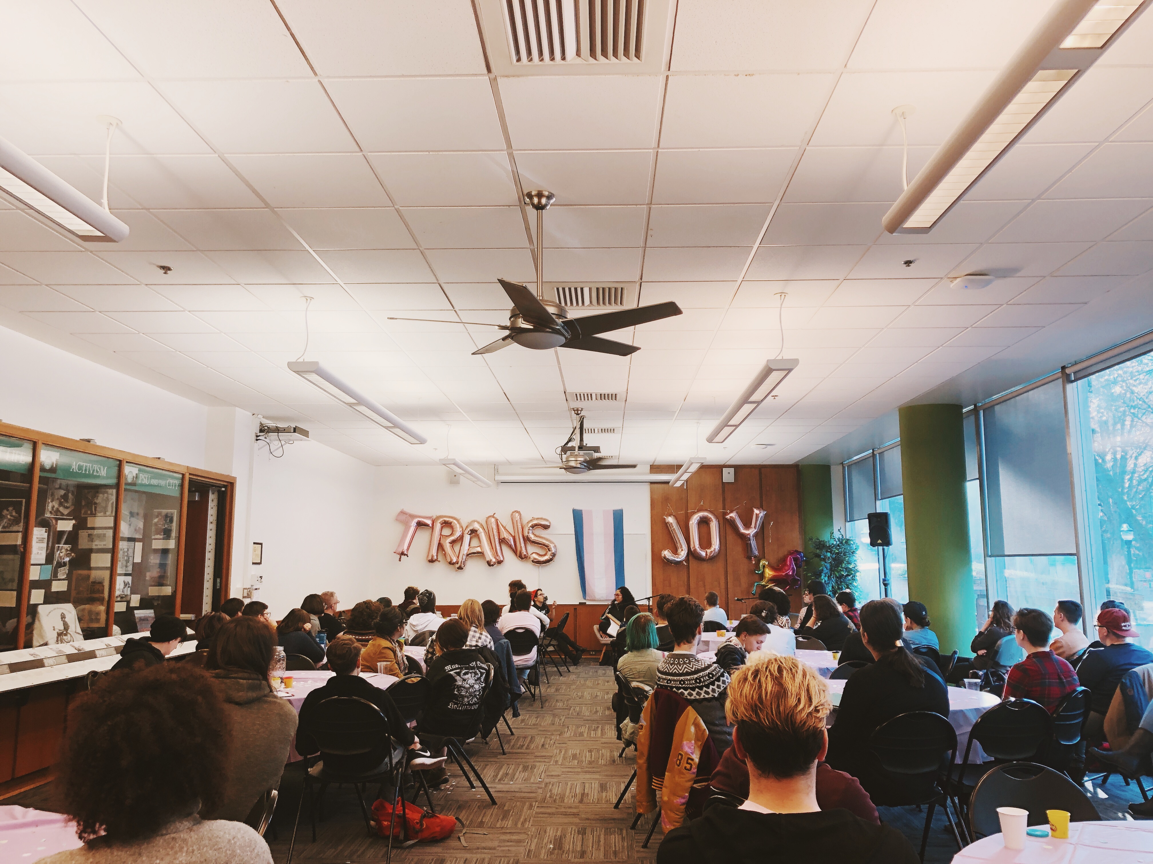 trans student sit around tables towards Tuck Woodstock from Gender Reveal podcast and Alyssa Pariah, local TWOC activist. pink balloons read "trans joy" with a trans flag between